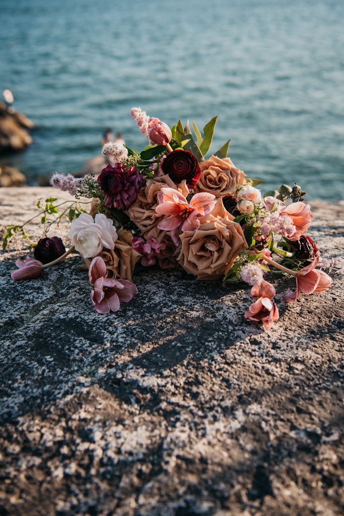 A rustic bouquet of assorted flowers lies on a stone surface by a tranquil sea.